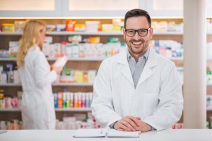 Portrait of a handsome pharmacist at the counter of a drugstore, female colleague working in the background.